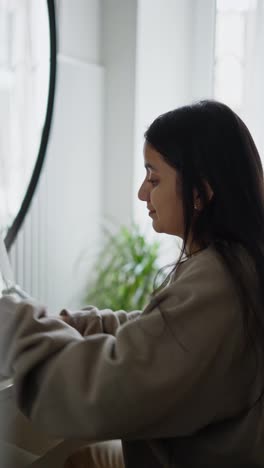 Vertical-video-of-a-confident-brunette-girl-in-a-gray-sweater-doing-Makeup-and-trying-the-smell-of-new-perfume-in-front-of-a-large-mirror-in-a-modern-apartment